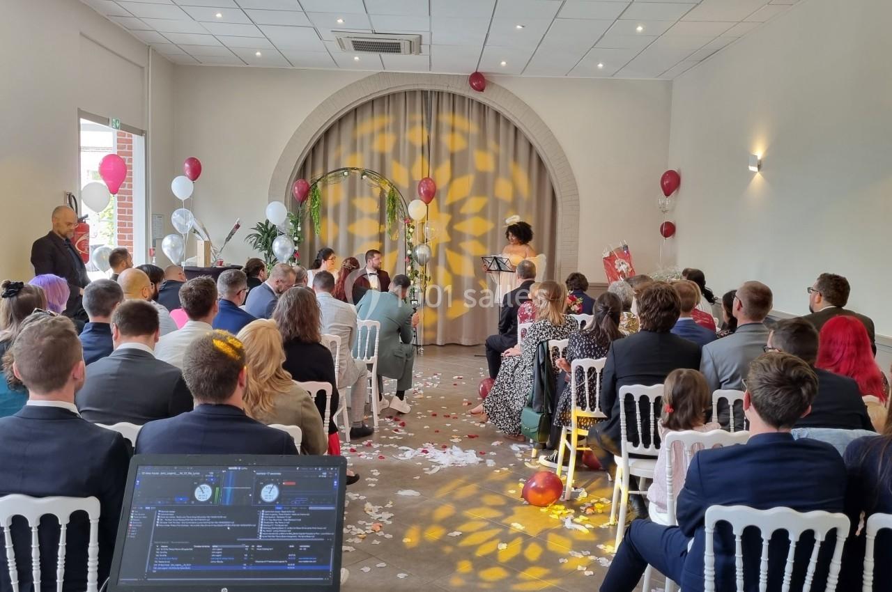 Salle de cérémonie avec invités assis, décorations festives et une personne lisant devant un couple sous une arche.