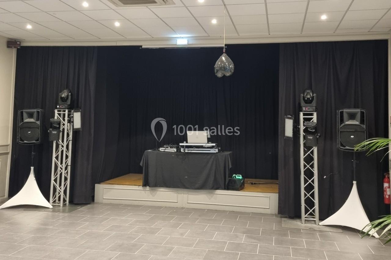 Scène avec rideaux noirs, table de mixage, enceintes sur pieds et éclairage suspendu dans une salle carrelée.