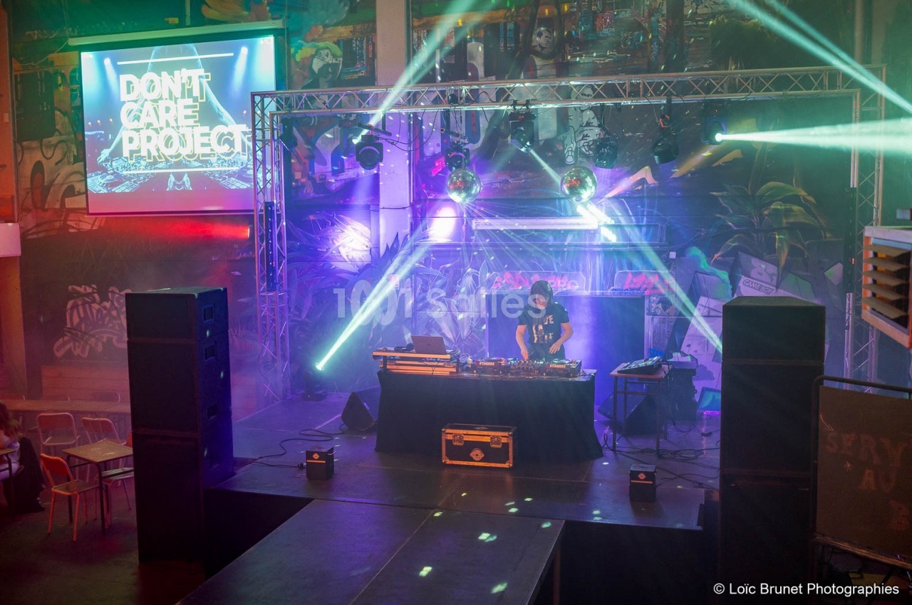 Un DJ mixe sur une scène éclairée par des projecteurs colorés, avec un écran affichant ’Don't Care Project’.