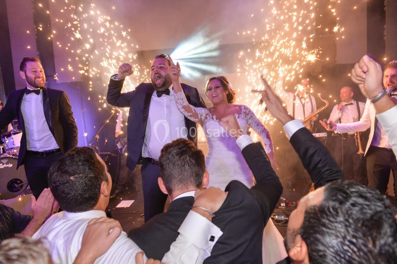 Un couple en tenue de mariage danse entouré d'invités en costume, avec des étincelles et un groupe en arrière-plan.