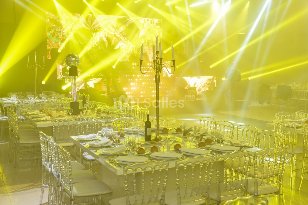 Salle de réception illuminée par des projecteurs jaunes, avec tables dressées et chaises transparentes.