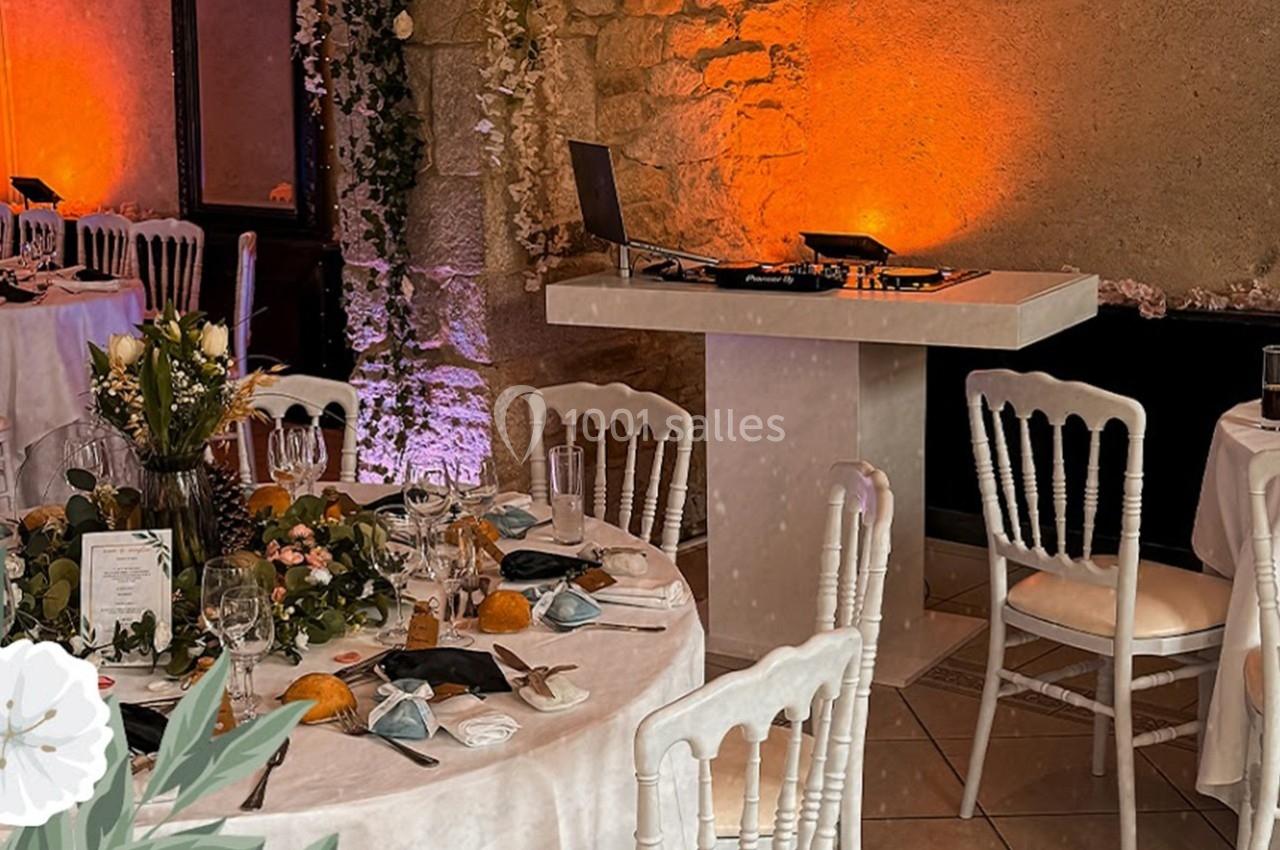 Salle de réception décorée avec des tables rondes dressées, des chaises blanches et une table de DJ en arrière-plan.