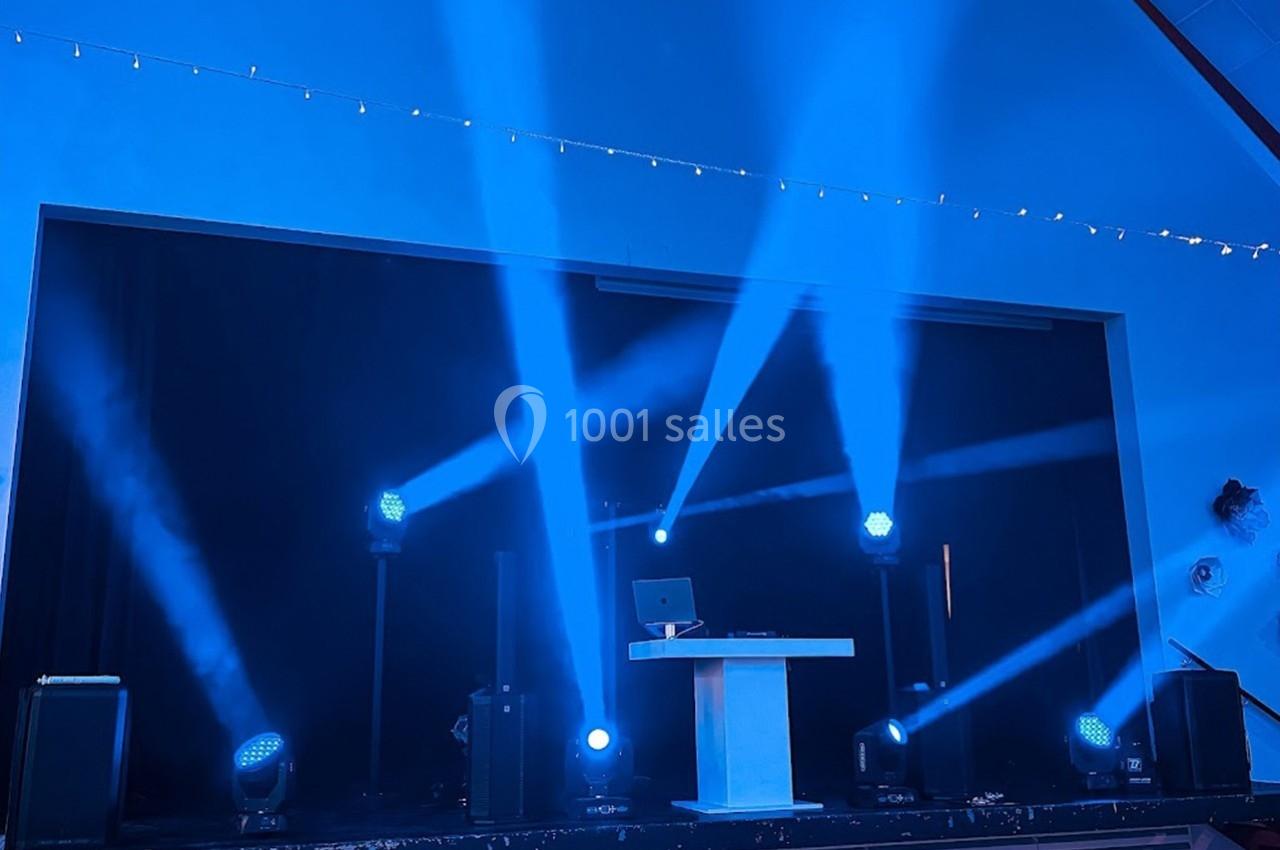 Scène éclairée par des projecteurs bleus avec une table blanche et un ordinateur au centre.
