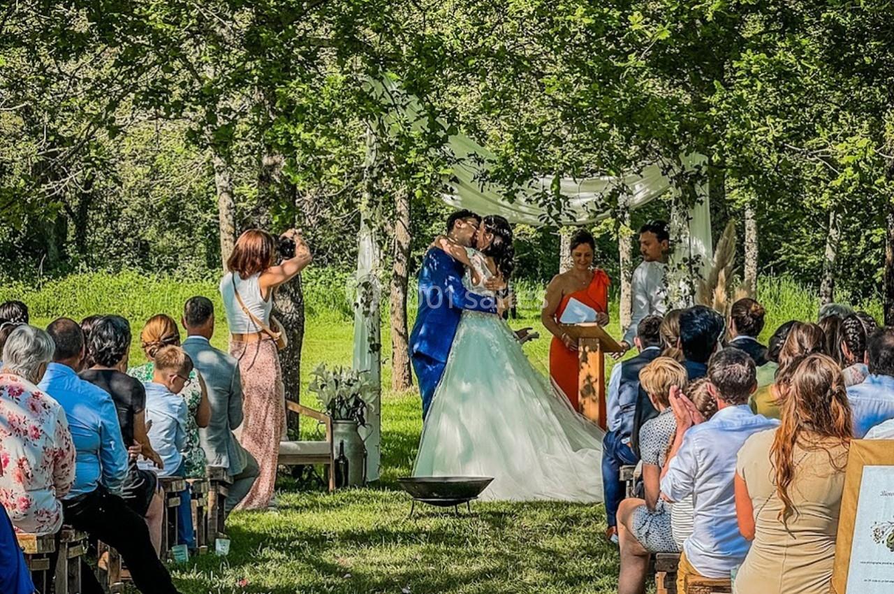 Un couple échange un baiser lors d'une cérémonie de mariage en plein air, entouré d'invités assis sous des arbres.