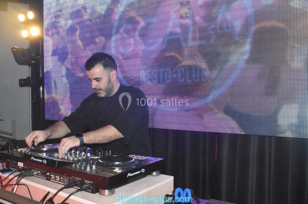 Un DJ mixe de la musique sur une table de mixage devant un écran lumineux affichant des motifs colorés.