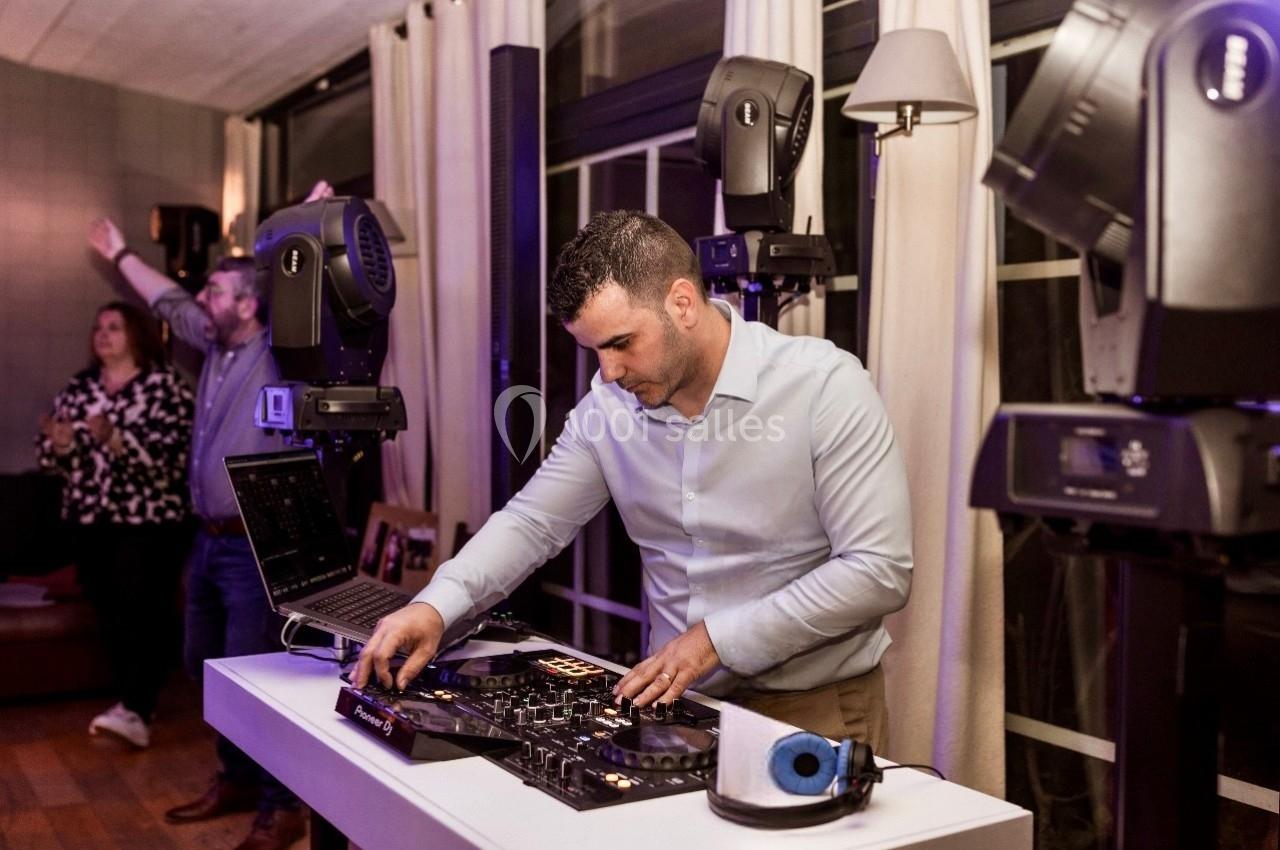 Un DJ mixe de la musique sur une table de mixage dans une salle éclairée, avec des invités en arrière-plan.