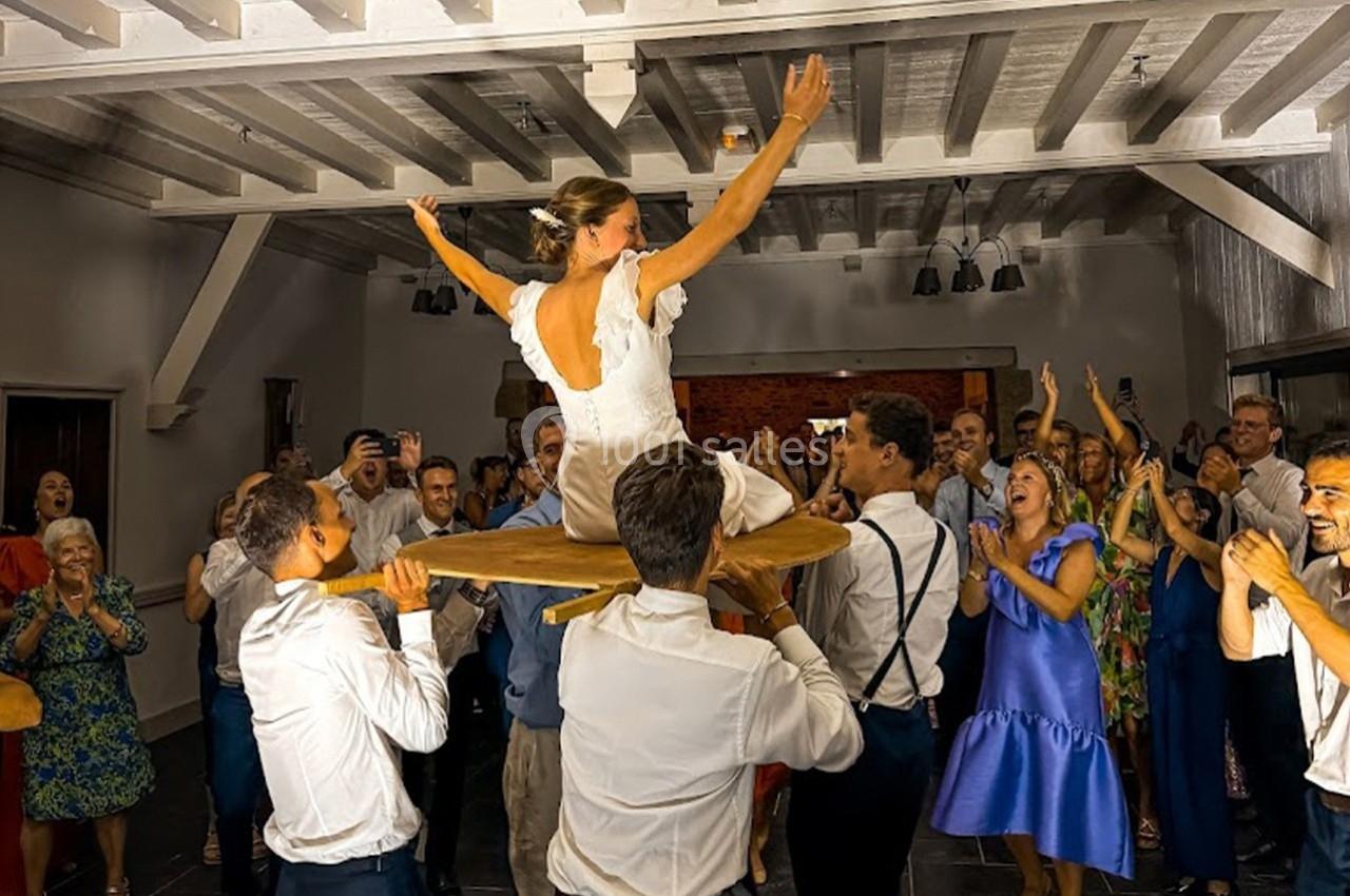 Une mariée est soulevée sur une chaise par des invités lors d'une fête dans une salle animée.