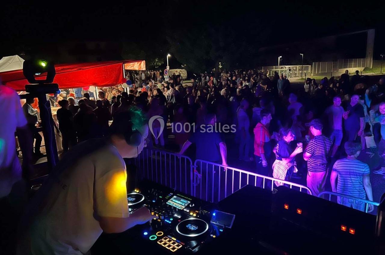 Un DJ mixe sur une scène en plein air face à une foule dansante sous des éclairages colorés, de nuit.