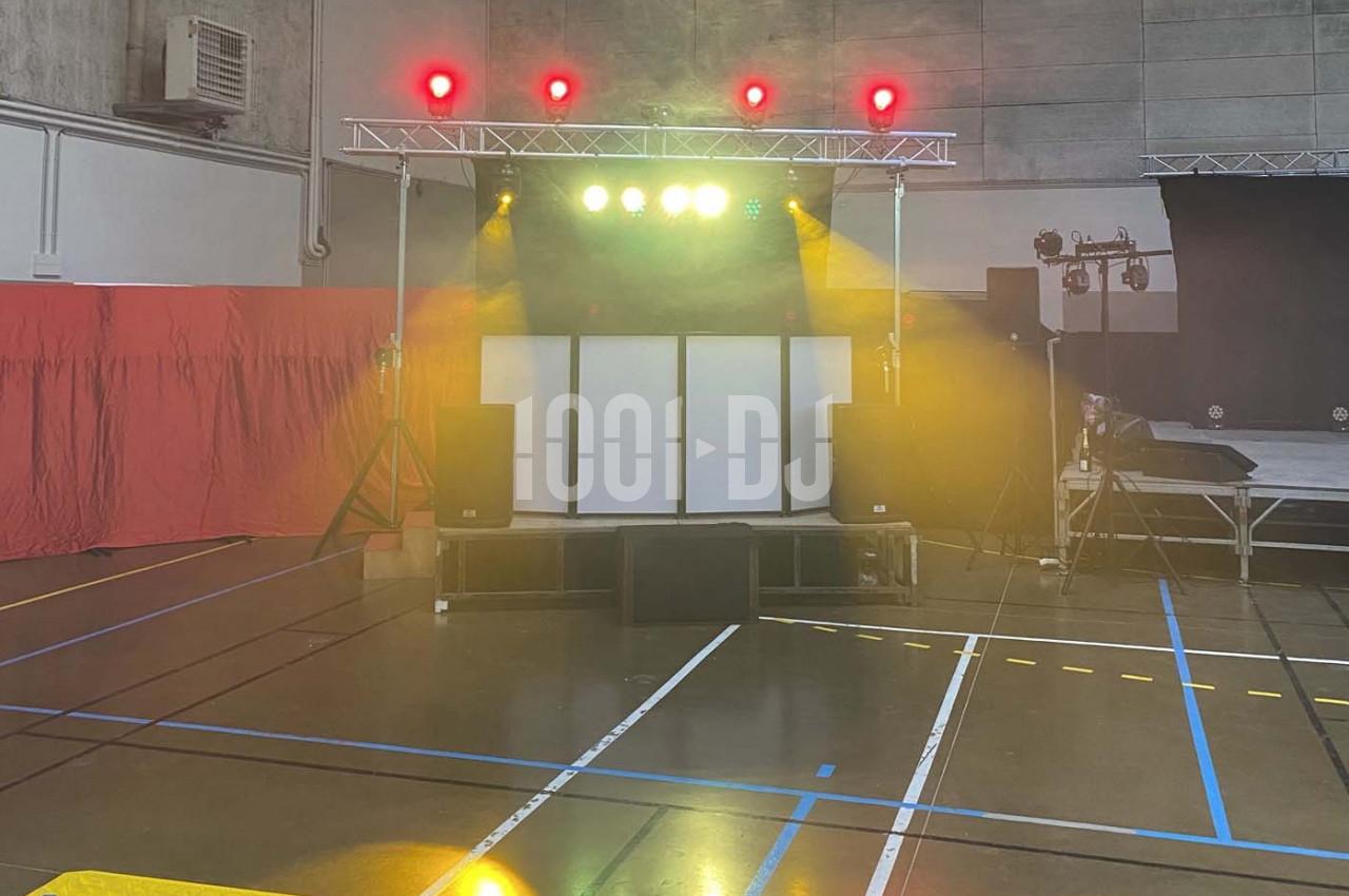 Scène avec éclairage coloré et équipement audio installée dans un gymnase aux murs gris et sol marqué de lignes.
