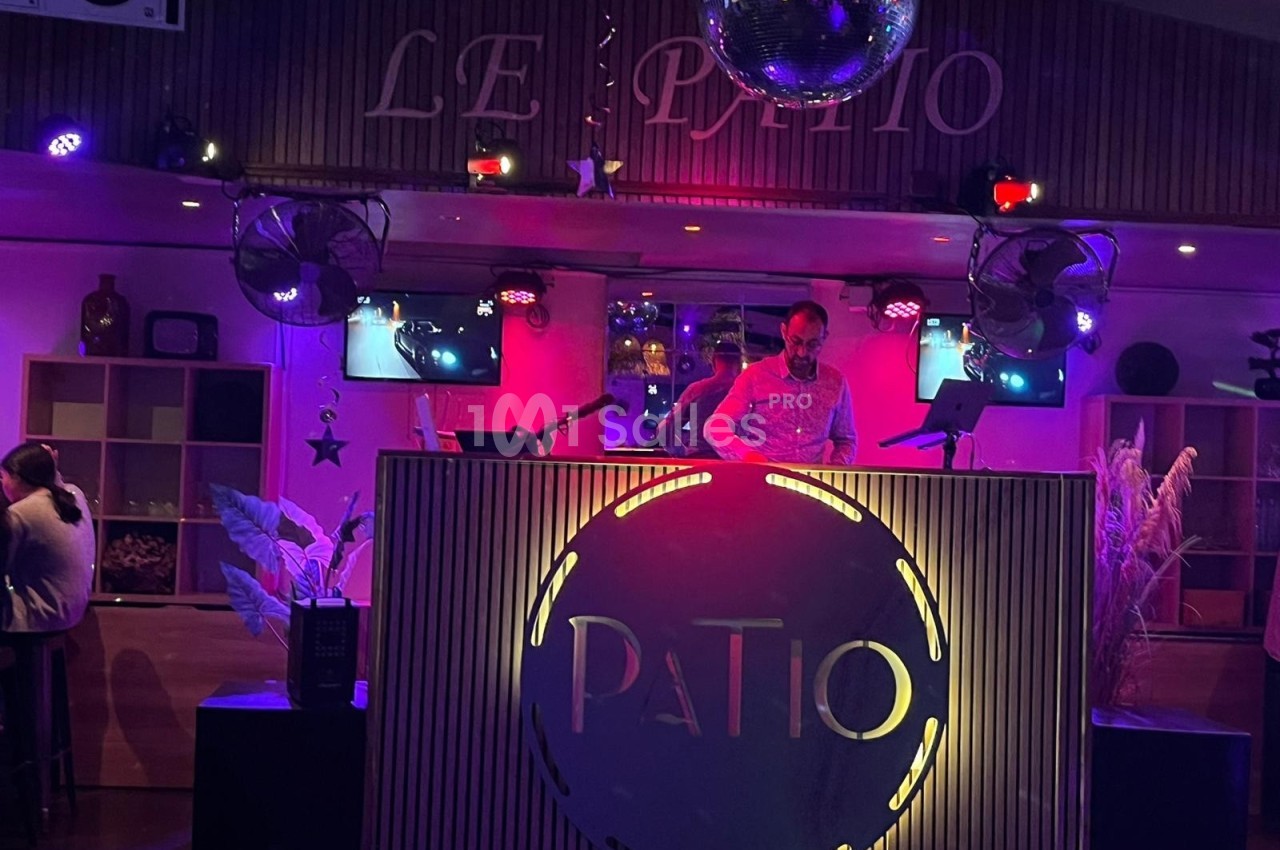 Un DJ mixe sur une scène éclairée par des lumières colorées dans un lieu nommé ’Le Patio’.