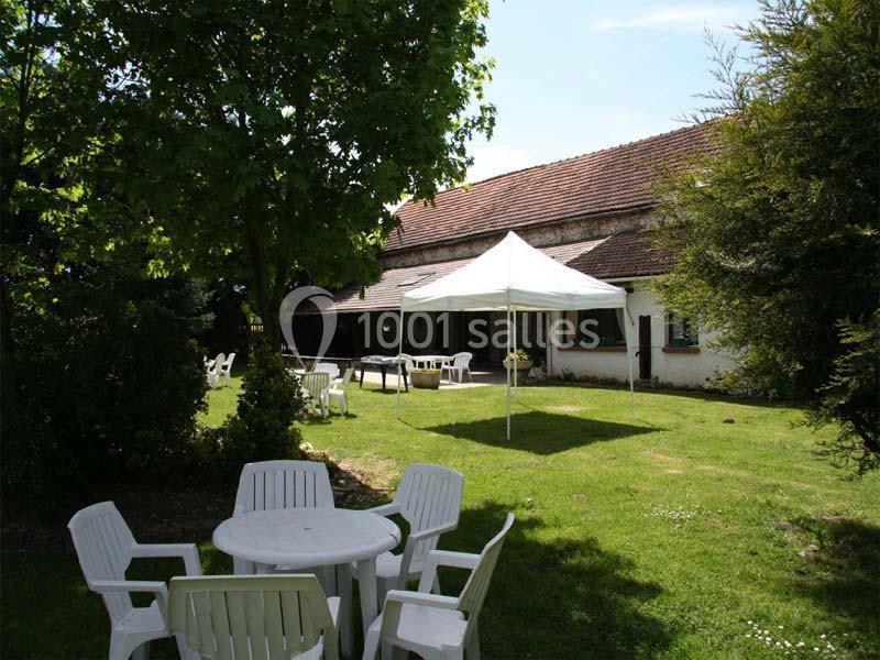 Jardin avec table et chaises blanches en plastique, tonnelle blanche, arbres et maison en arrière-plan.