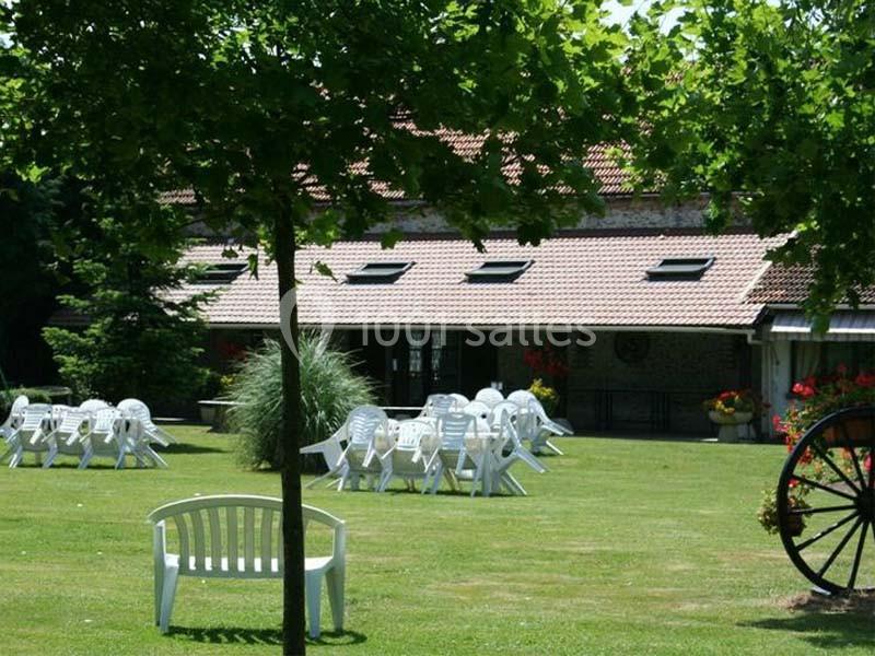 Jardin avec pelouse, chaises blanches disposées autour de tables, arbres et bâtiment en arrière-plan.