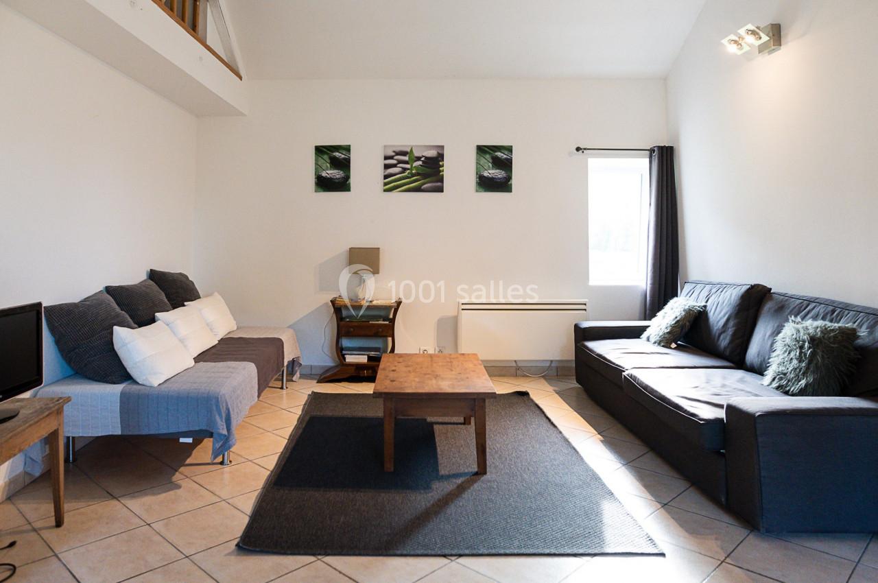 Salon lumineux avec canapé, fauteuil, table basse en bois, tapis gris et décoration murale, éclairé par une fenêtre.