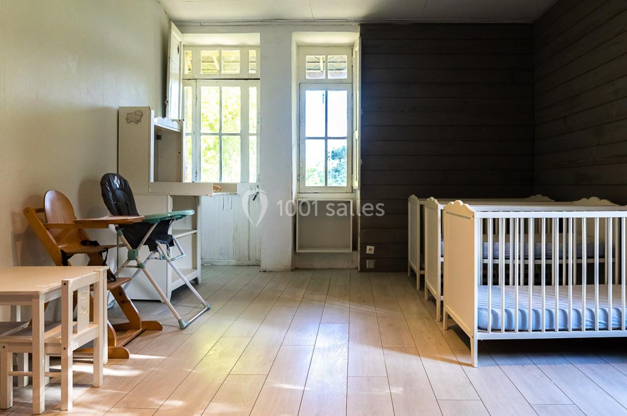 Chambre lumineuse avec deux lits pour bébé, une chaise haute, une table et des murs en bois et en blanc.