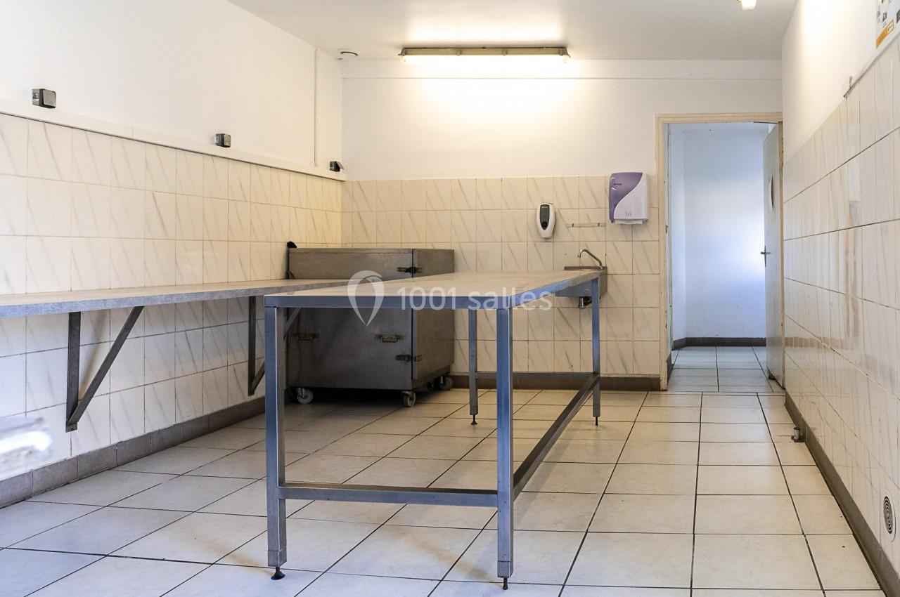 Salle carrelée avec deux grandes tables en inox, un évier mural et un éclairage fluorescent au plafond.