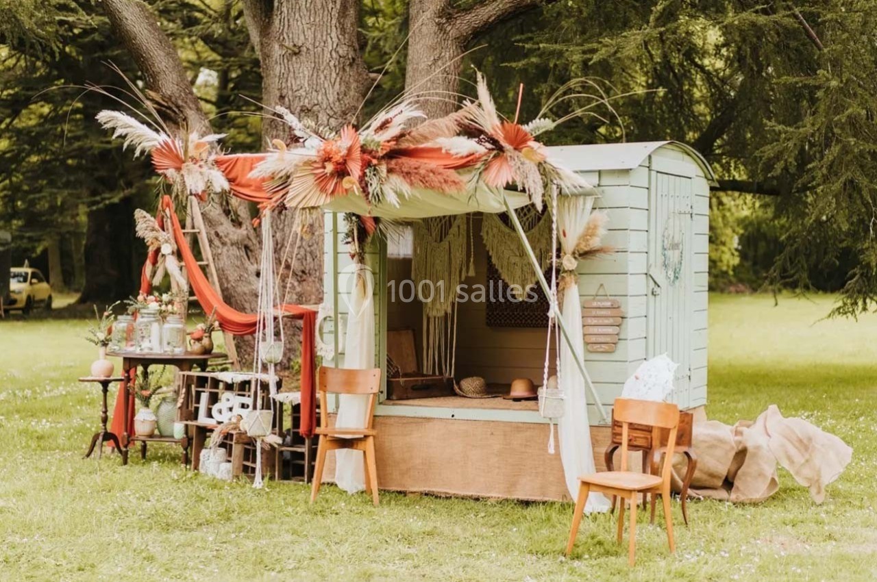Abri en bois décoré de fleurs séchées et tissus rouges, entouré de chaises et d'objets sur une pelouse verdoyante.
