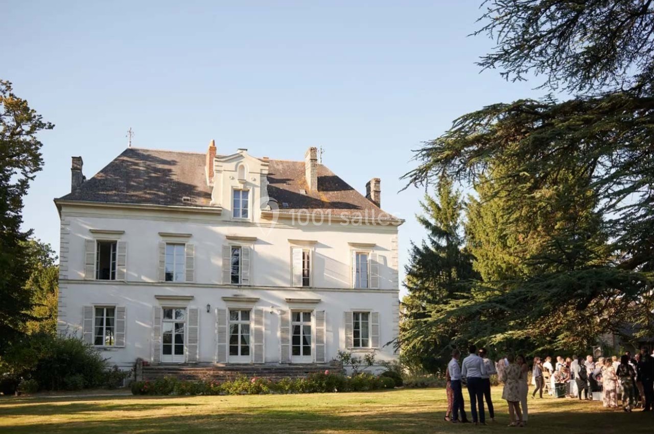 Façade d'un manoir blanc entouré d'un jardin, avec un groupe de personnes rassemblées à l'ombre des arbres.