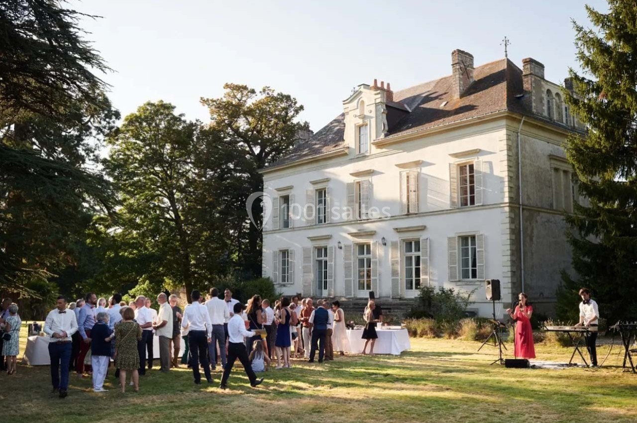 Groupe de personnes rassemblées dans un jardin devant une grande maison ancienne, avec des musiciens jouant en extérieur.