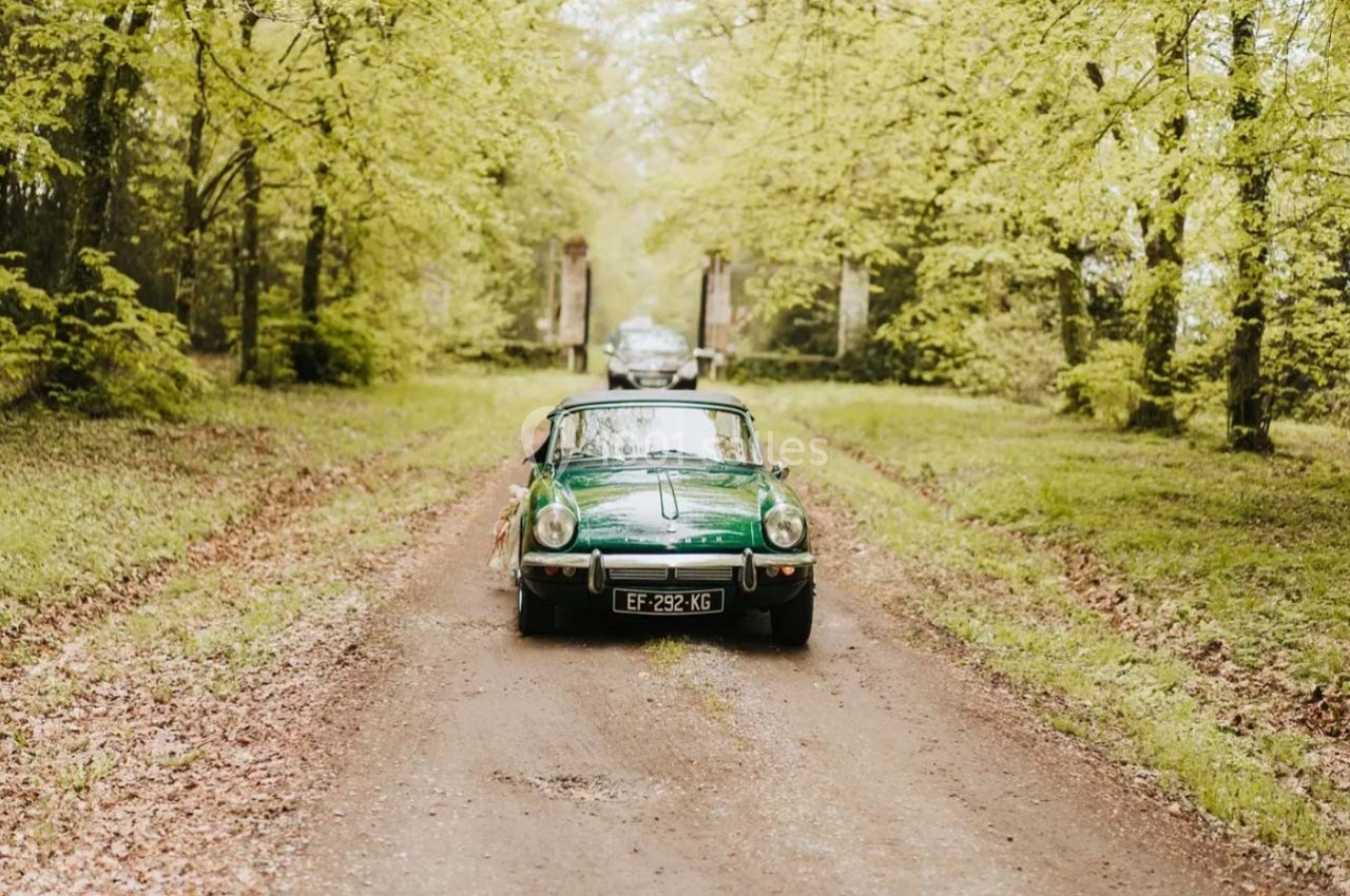 Voiture ancienne verte sur un chemin forestier bordé d'arbres, avec une autre voiture en arrière-plan.