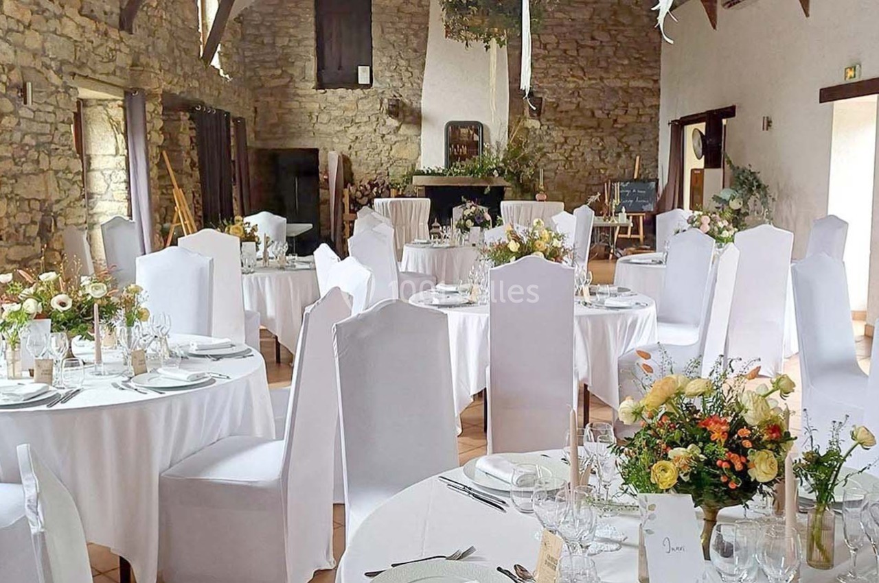 Salle de réception en pierre avec tables rondes dressées, nappes blanches et décorations florales.