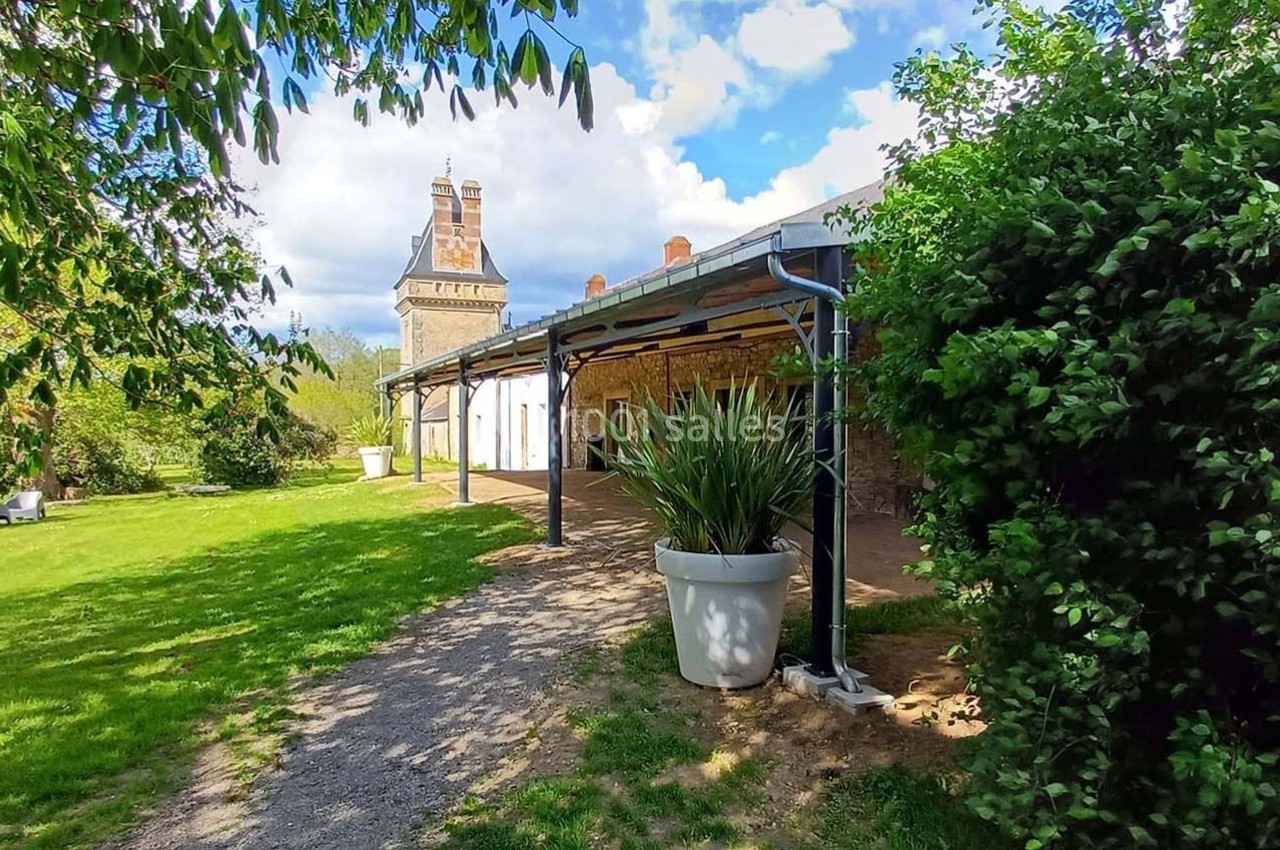 Allée ombragée longeant un bâtiment en pierre avec une tour en arrière-plan, entourée de verdure.