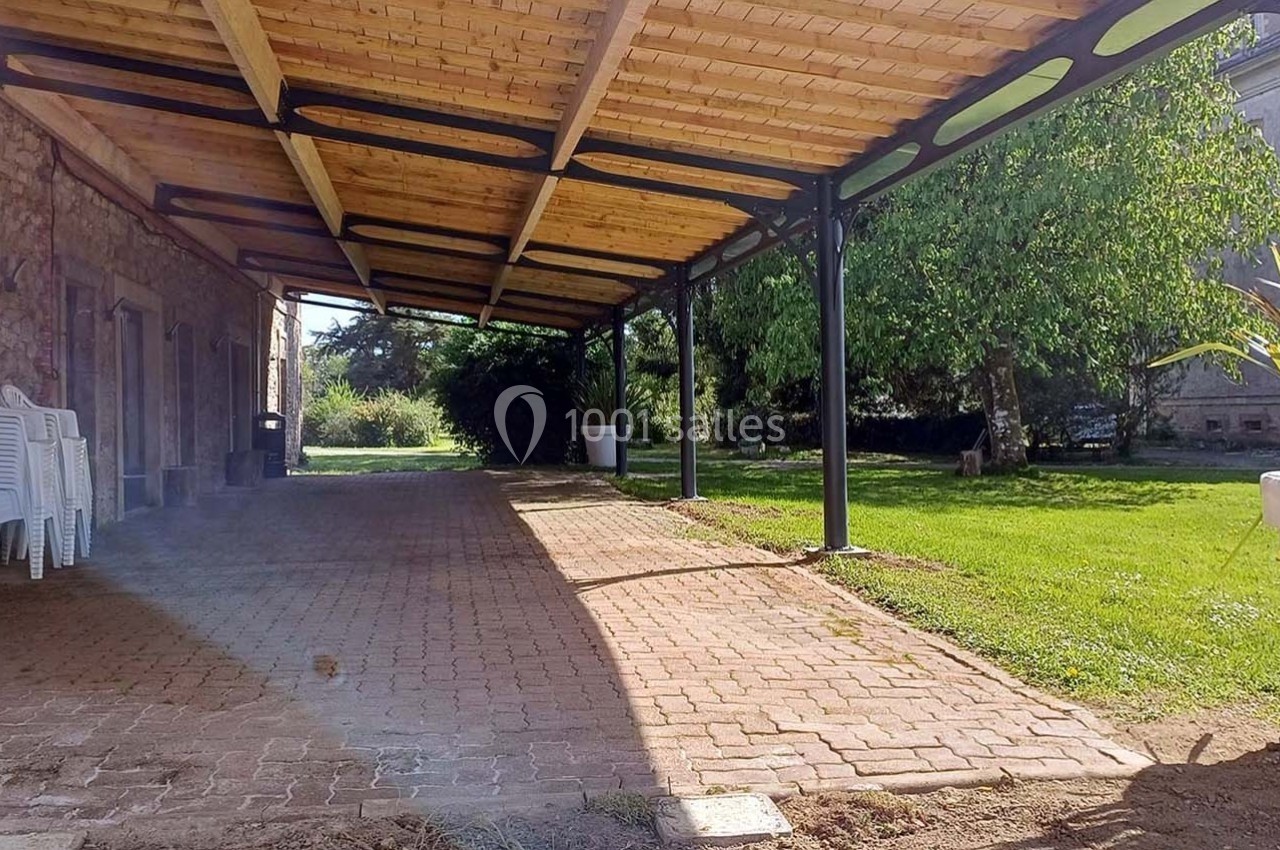 Terrasse couverte avec structure en bois et métal, bordée d'un espace vert et d'un bâtiment en pierre.