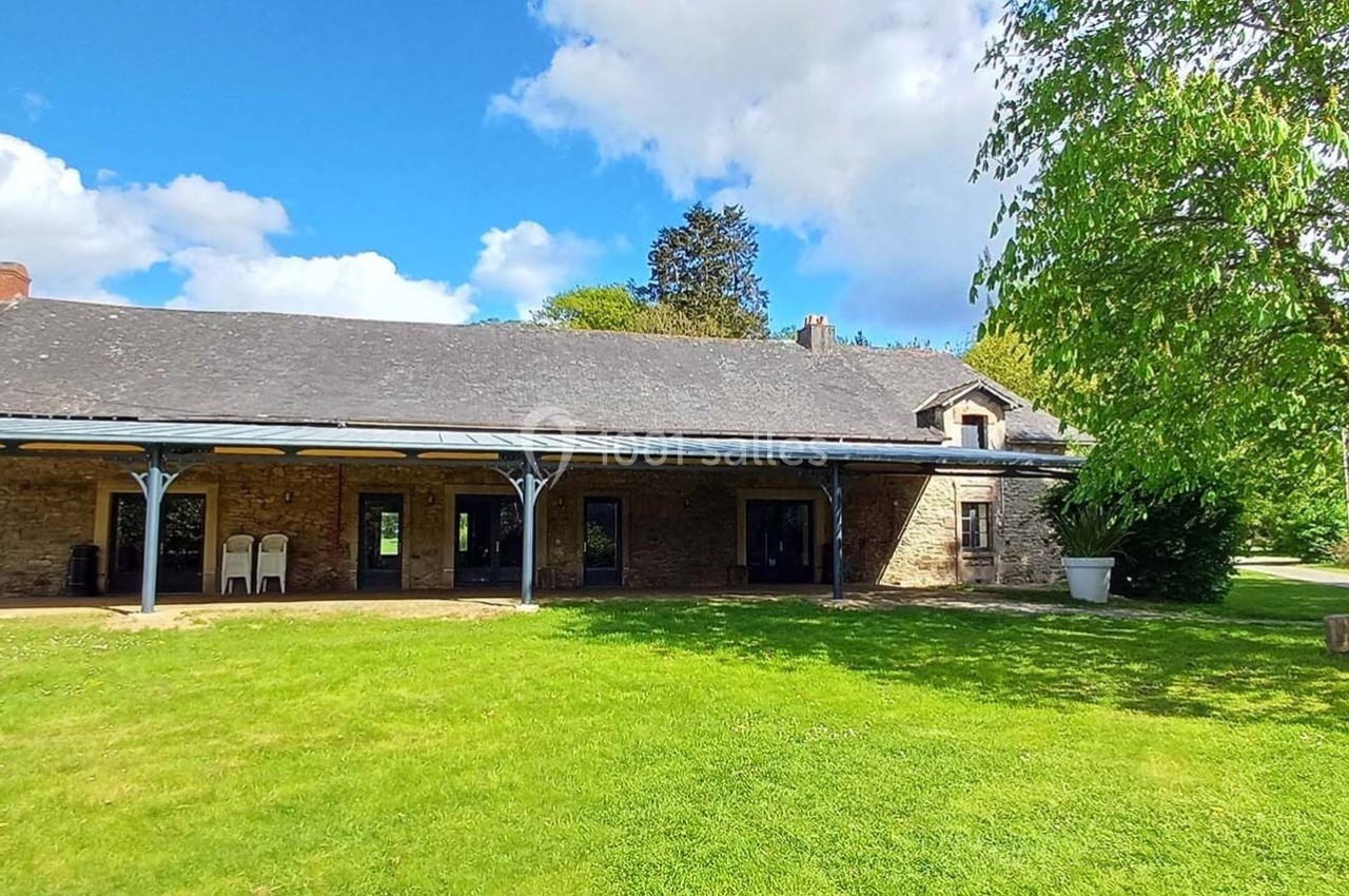 Bâtiment en pierre avec toit en ardoise, terrasse couverte et pelouse verdoyante sous un ciel partiellement nuageux.