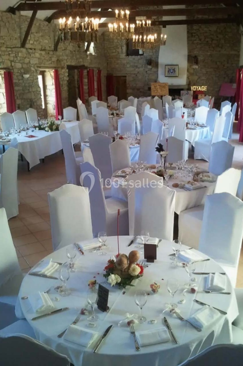 Salle de réception avec tables rondes dressées, nappes blanches et chaises couvertes, dans un cadre en pierre apparente.