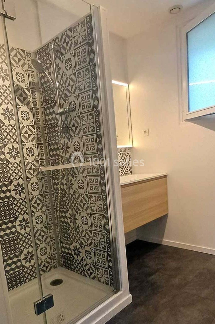 Douche avec paroi vitrée, carrelage à motifs géométriques noirs et blancs, meuble vasque et fenêtre en arrière-plan.