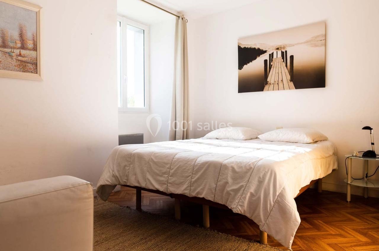 Chambre lumineuse avec un lit double, parquet en bois, tableau mural et fenêtre laissant entrer la lumière naturelle.