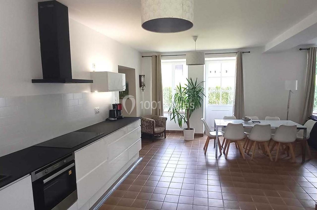 Cuisine ouverte moderne avec îlot, coin repas, table blanche et chaises, éclairée par de grandes fenêtres.