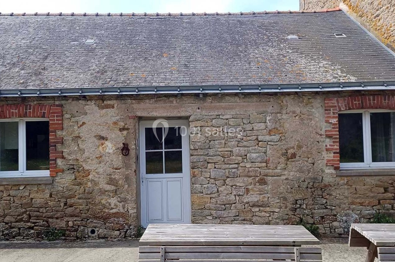 Façade en pierre d'une petite maison avec une porte blanche et deux fenêtres, entourée de bancs en bois.