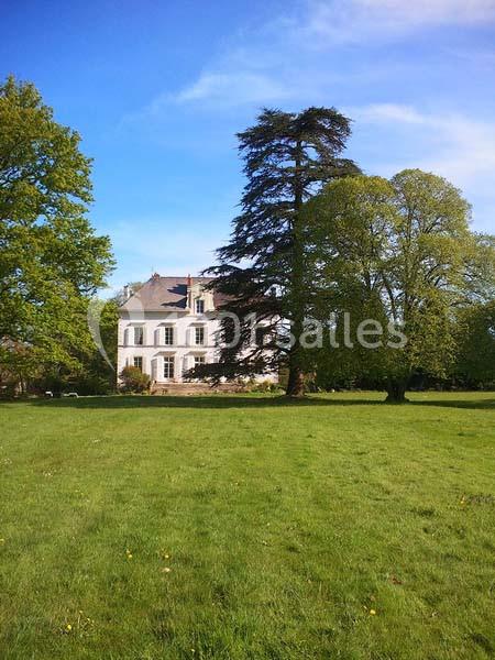 Manoir blanc entouré d'arbres, situé dans un grand parc verdoyant sous un ciel bleu.