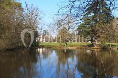 Étang calme entouré d'arbres avec un bâtiment blanc visible en arrière-plan sous un ciel bleu.