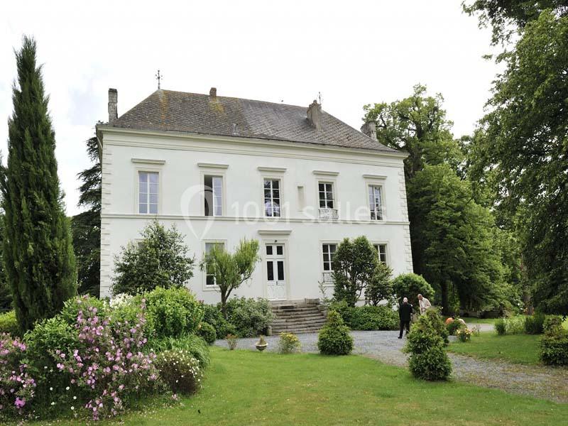 Grande maison blanche entourée d'un jardin verdoyant avec des arbres, arbustes et fleurs, deux personnes au premier plan.