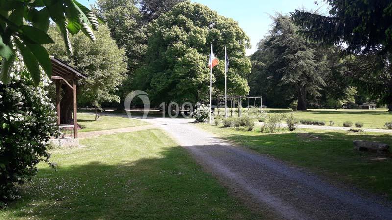 Allée gravillonnée traversant un parc arboré avec un drapeau et des structures de jeux en arrière-plan.