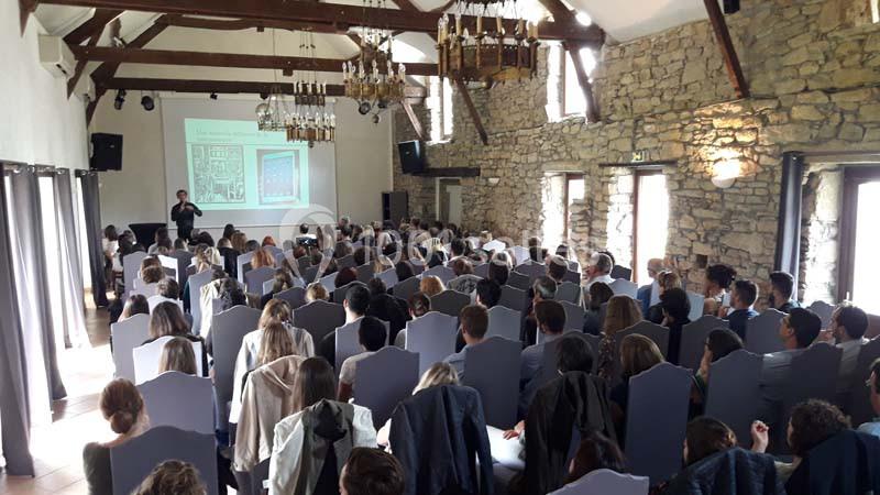 Salle en pierre avec poutres apparentes, remplie de personnes assises assistant à une présentation projetée.
