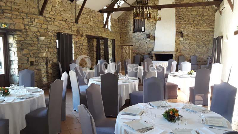 Salle de réception en pierre avec tables rondes dressées, chaises grises et décoration florale.