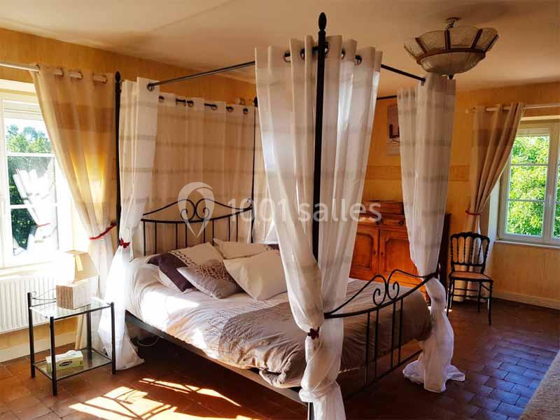 Lit à baldaquin avec voilages blancs dans une chambre lumineuse aux murs clairs et sol en terre cuite.