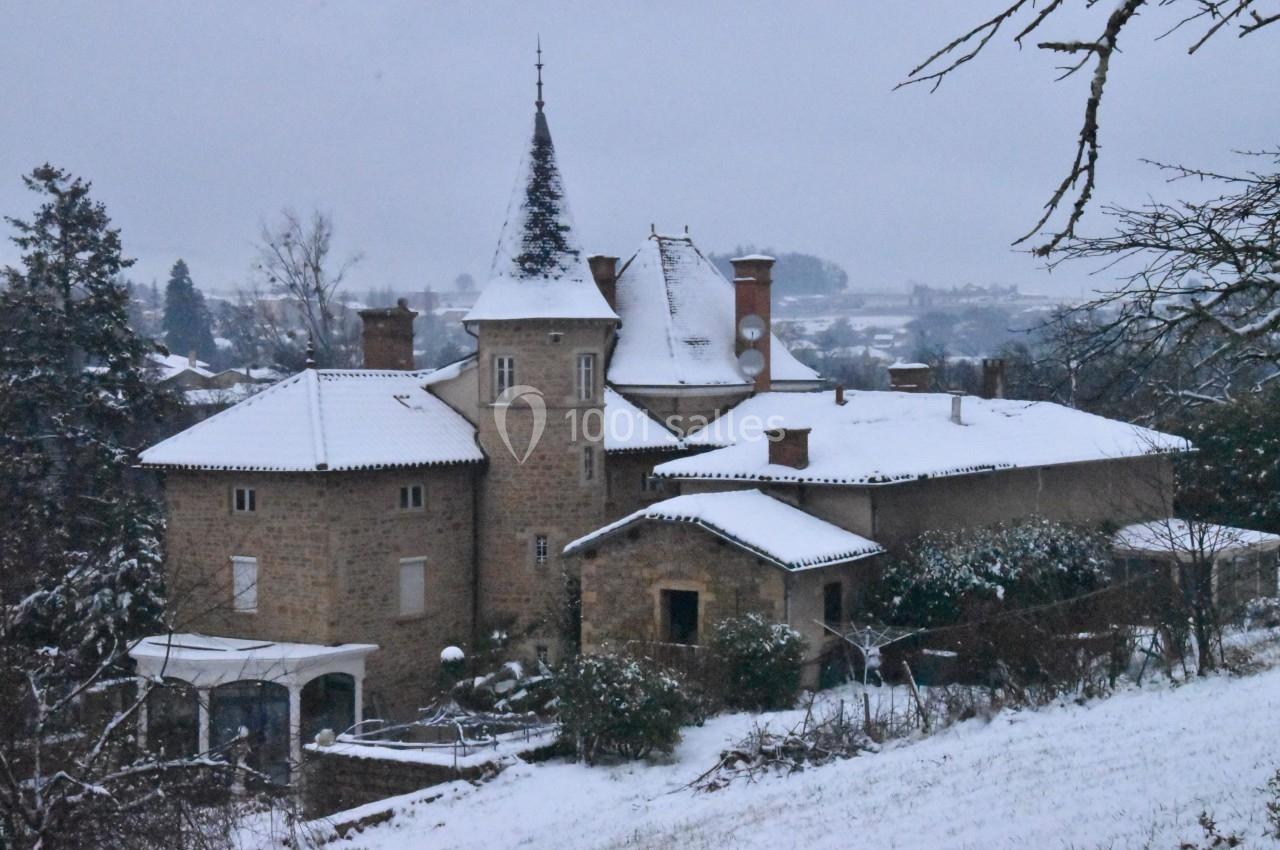 Manoir en pierre avec toit enneigé, entouré d'arbres et de végétation sous un ciel gris d'hiver.