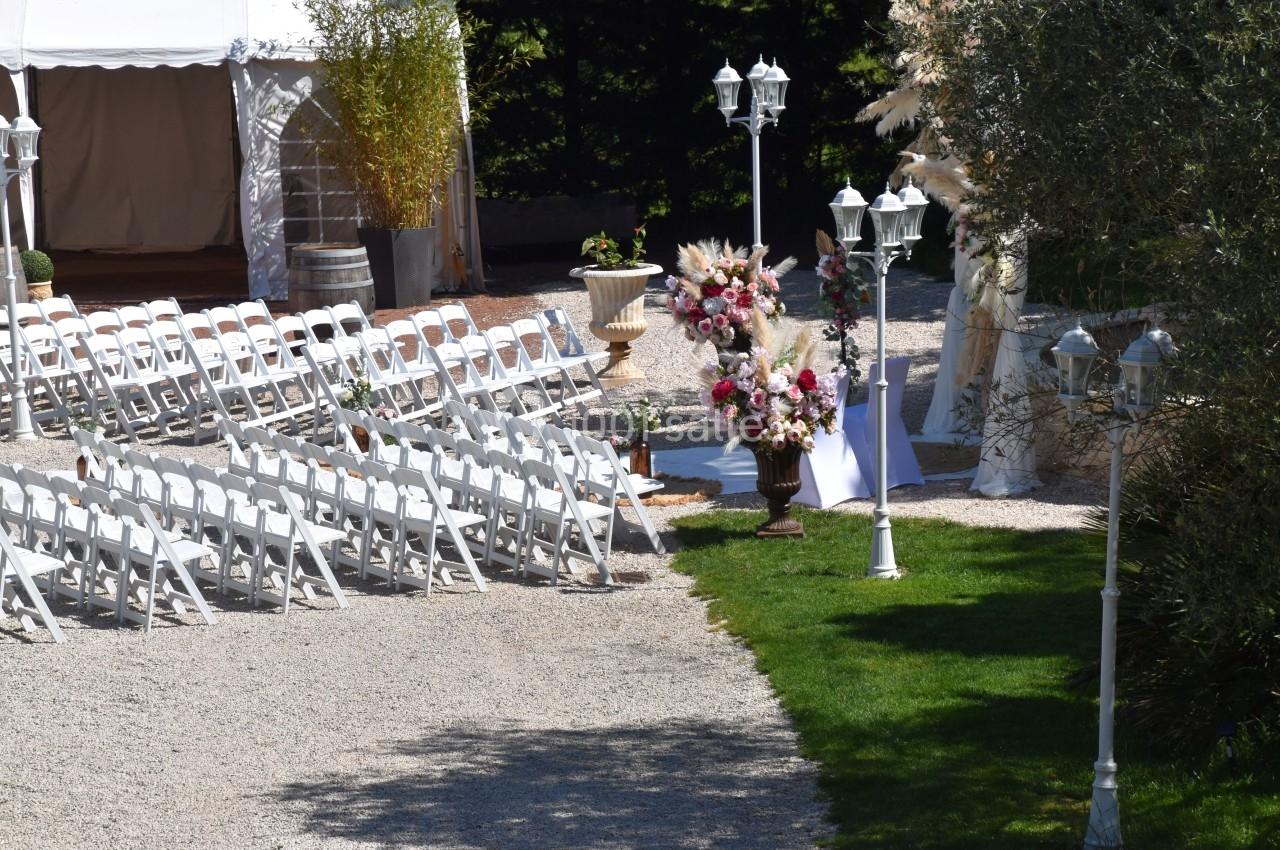 Chaises blanches alignées en extérieur pour une cérémonie, avec décorations florales et tente en arrière-plan.