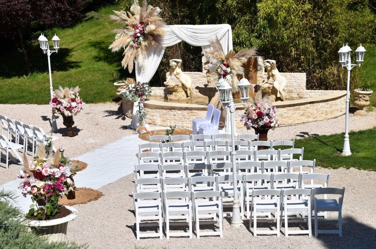 Cérémonie en extérieur avec chaises blanches alignées, arche décorée de fleurs et statues en arrière-plan.