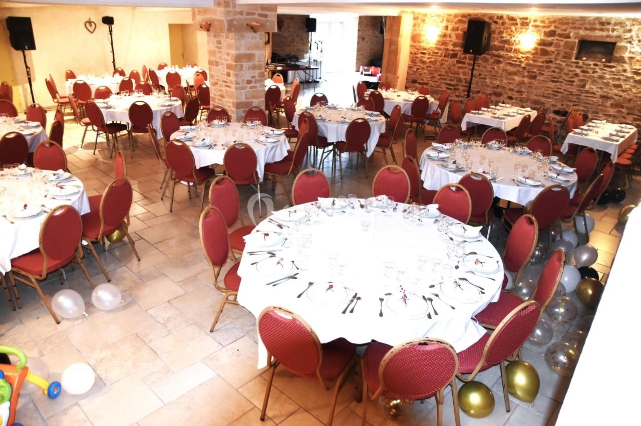 Salle de réception avec des tables rondes dressées, chaises rouges et décorations, dans un cadre en pierre apparente.