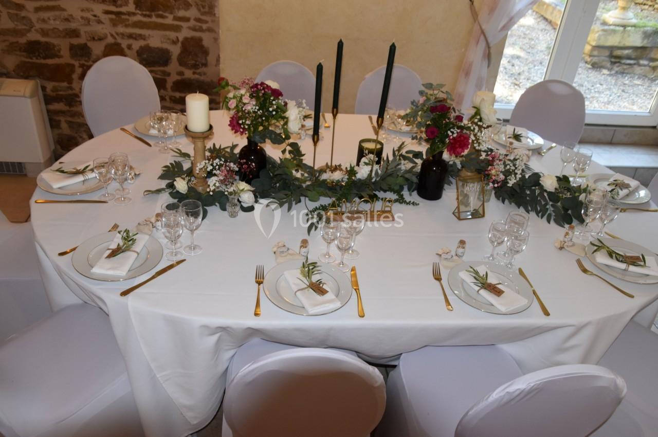 Table ronde élégamment dressée avec nappes blanches, bougies, fleurs et couverts dorés dans une salle en pierre.