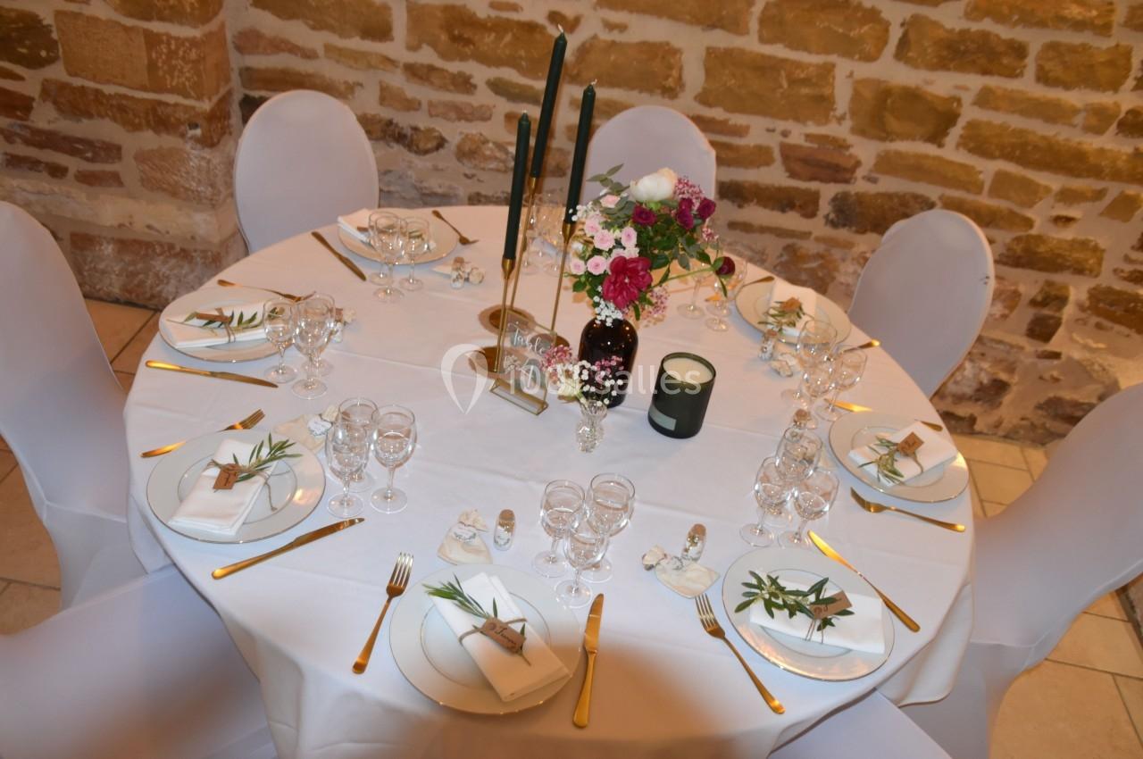 Table ronde décorée pour un repas, avec nappes blanches, bougies, fleurs et couverts dorés.
