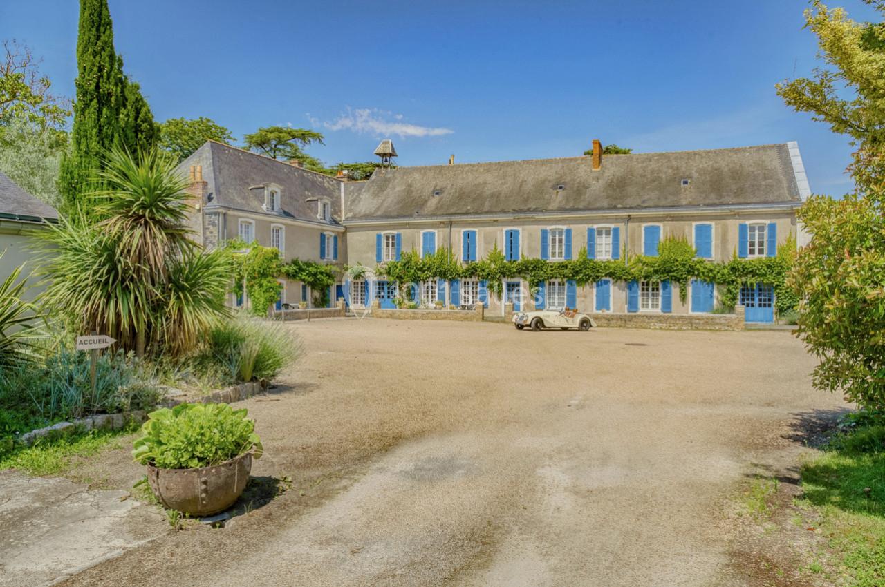 Cour d'une grande maison en pierre avec volets bleus, entourée de végétation et d'un ciel dégagé.