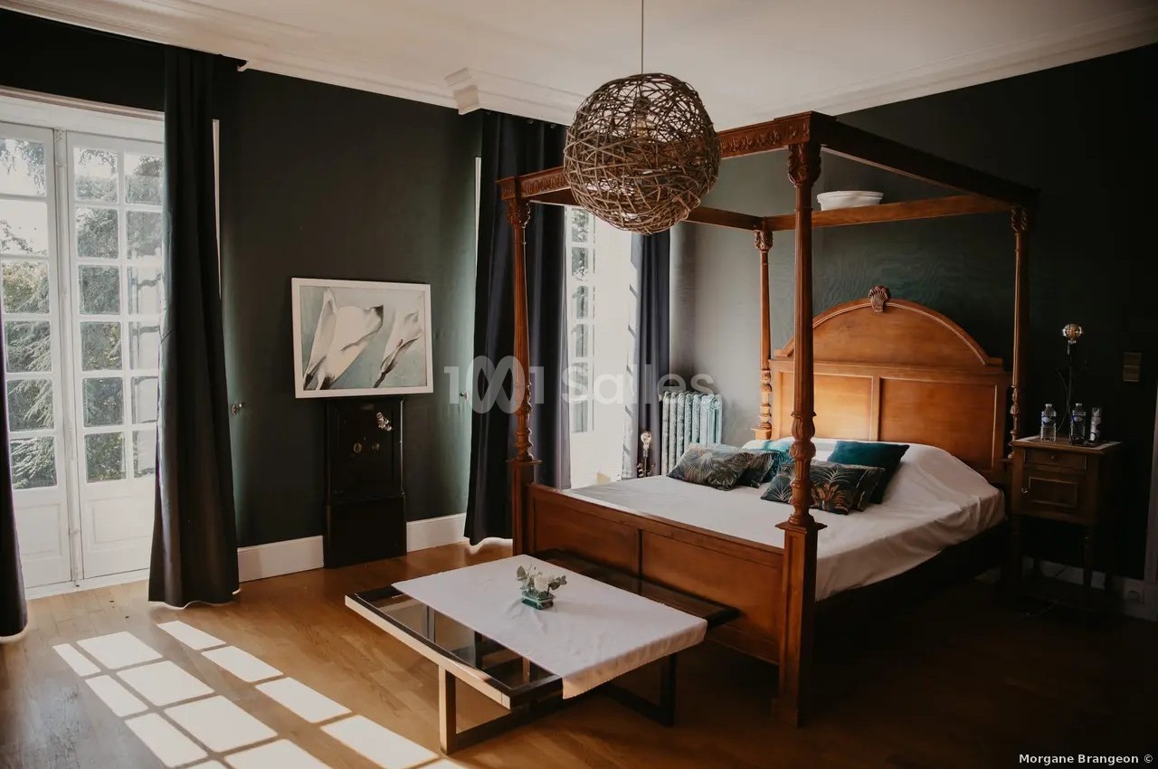 Chambre lumineuse avec lit à baldaquin en bois, table basse blanche et murs sombres, éclairée par une grande fenêtre.