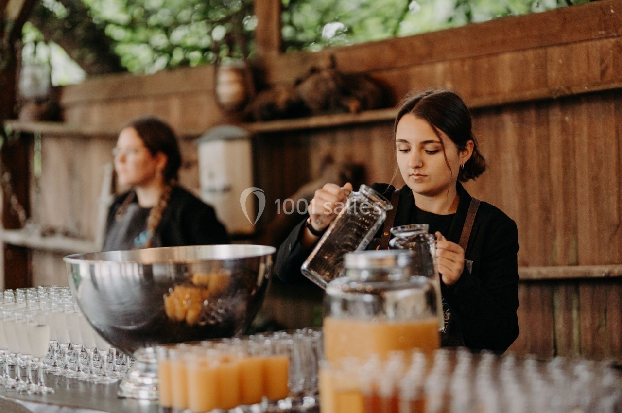 Une femme verse une boisson dans un verre sur un comptoir rempli de verres et de carafes, en extérieur.
