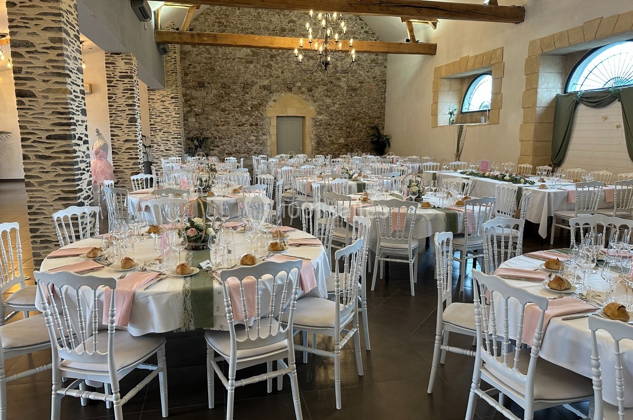 Salle de réception décorée avec des tables rondes dressées, nappes blanches et accents roses, sous un plafond en poutres…