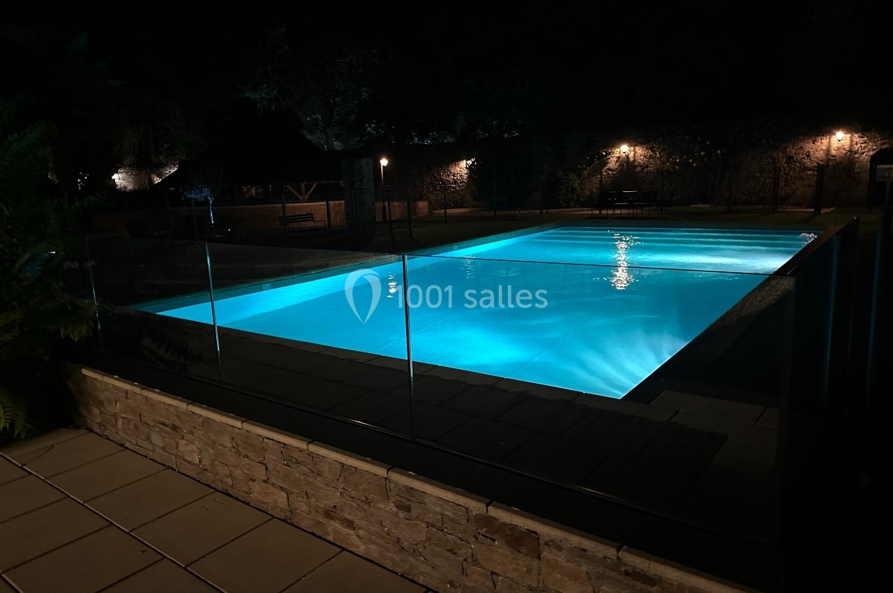 Piscine éclairée de nuit avec eau bleutée, entourée de dalles, d'une clôture en verre et d'arbres en arrière-plan.